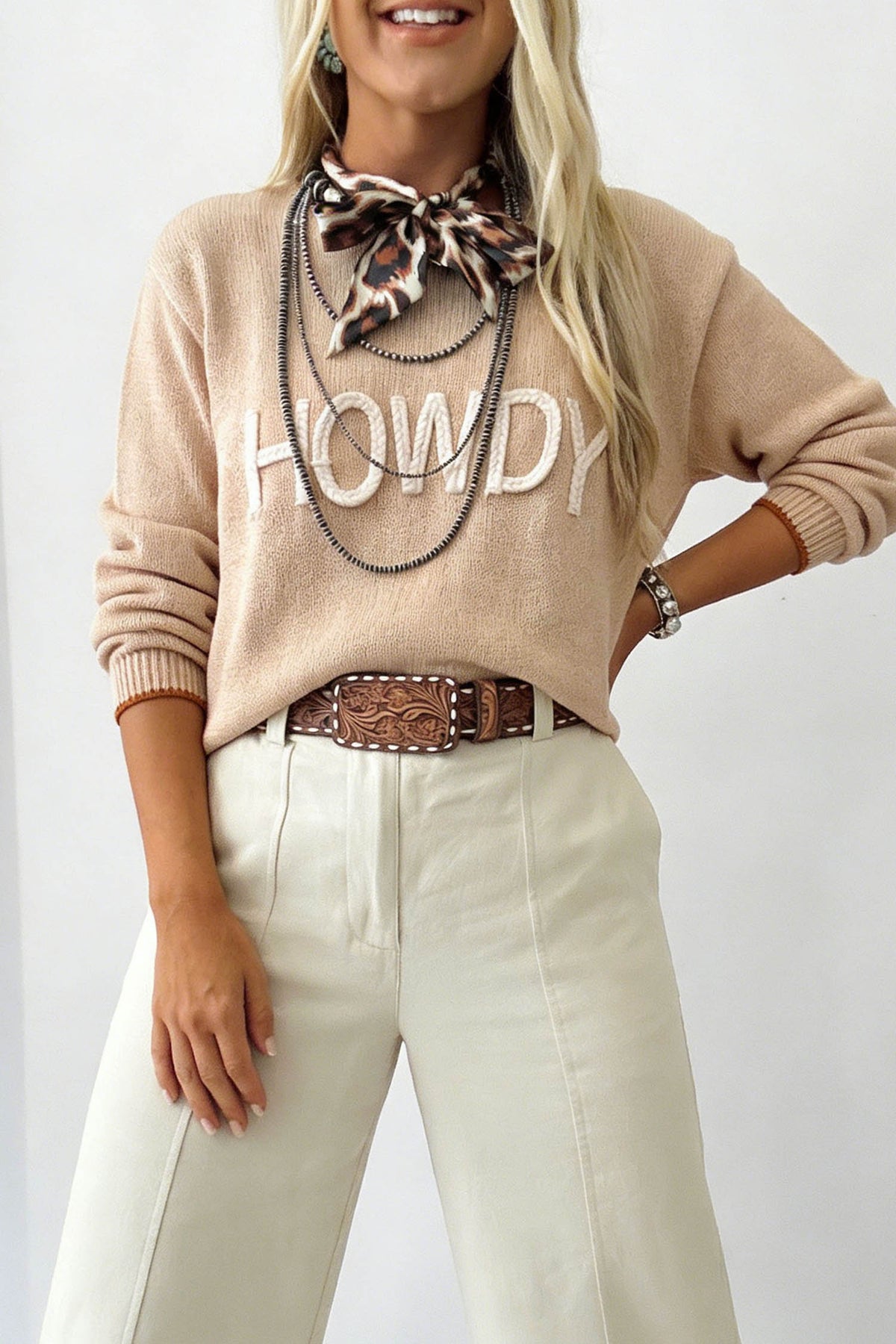 A woman wearing a beige crewneck sweater with 'HOWDY' in white varsity letters, styled with cream pants, a brown patterned belt, a leopard-print neck scarf, and layered silver necklaces.