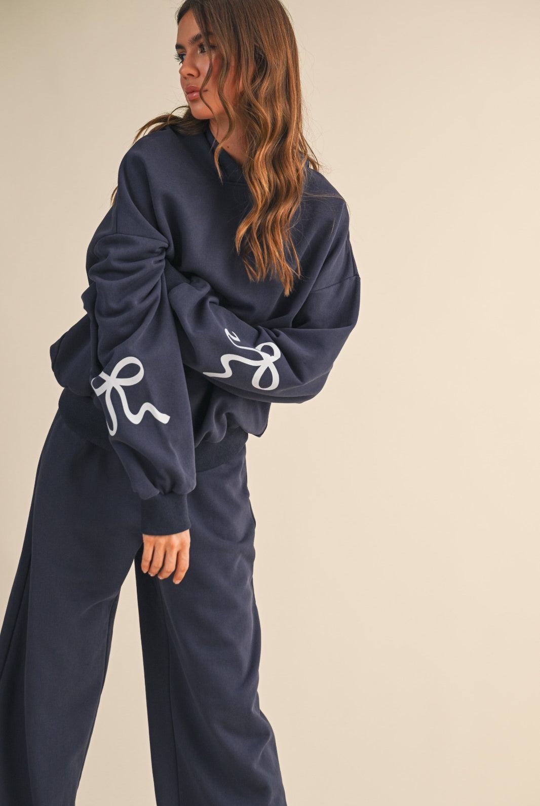 Woman wearing a navy blue hoodie sweatshirt with bow details on sleeves against a beige background
