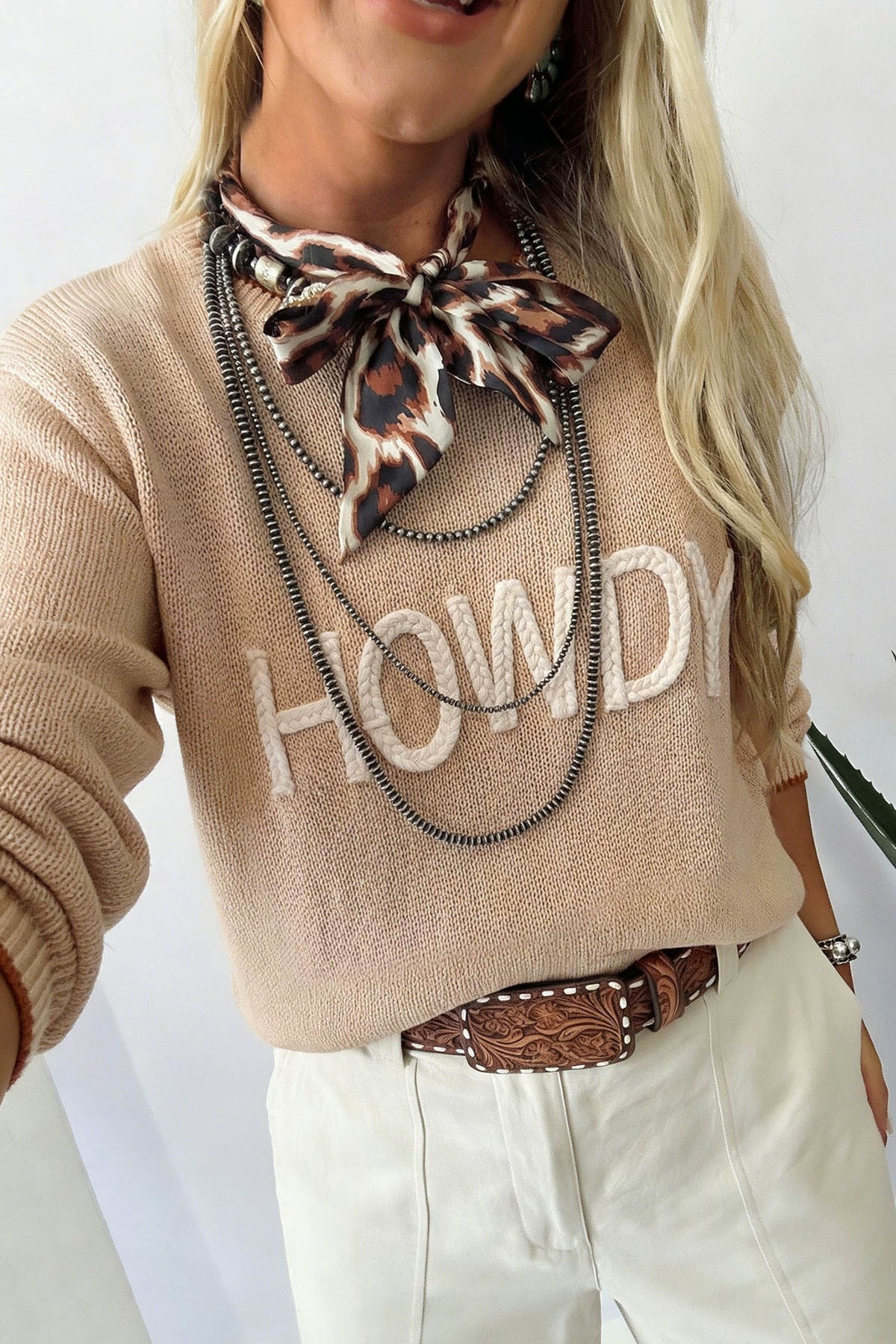 A woman wearing a beige knit sweater with 'HOWDY' in white letters across the chest, styled with a leopard-print neck scarf, layered black chains, a brown tooled belt, and off-white pants.