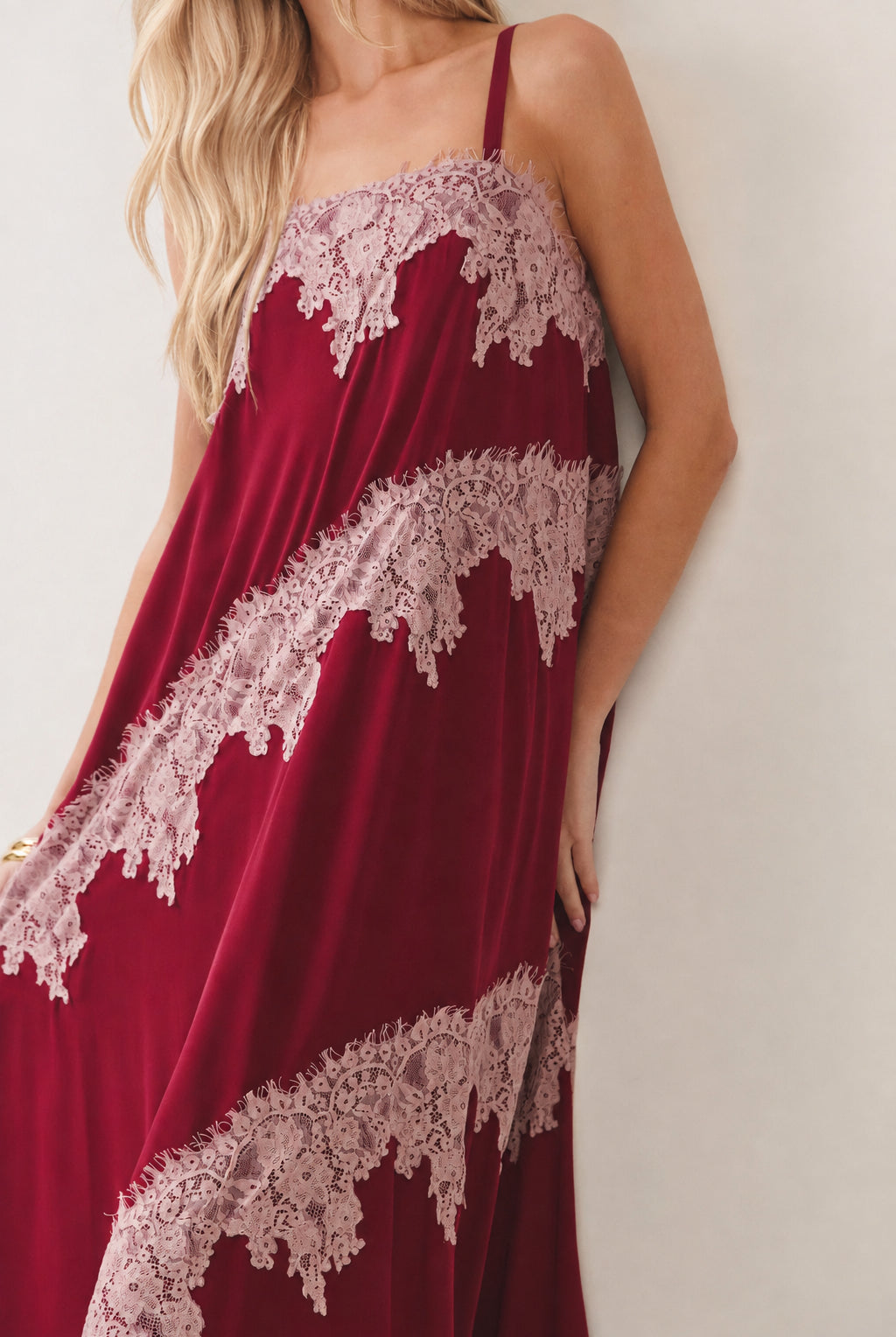 A woman wearing a burgundy satin slip dress with dusty pink lace trim, photographed against a plain off-white wall.