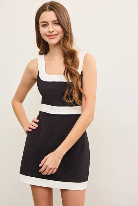 A young woman with long, wavy brown hair wears a black mini dress with a bold white trim along the square neckline, waistline, and hem. The dress has a structured, sleek fit, offering a stylish and modern look against a neutral background.
