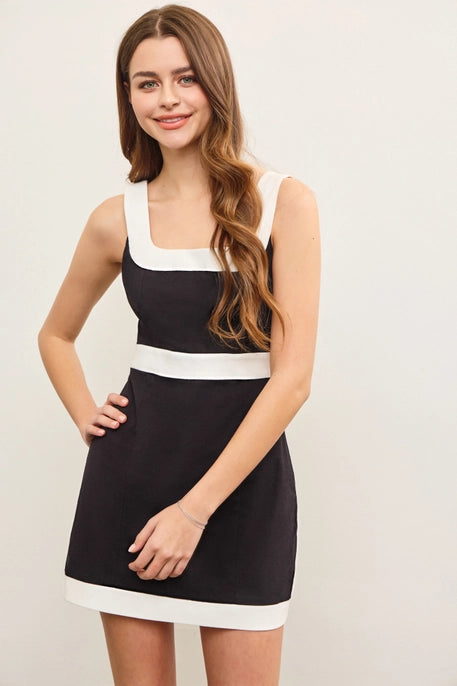 A young woman with long, wavy brown hair wears a black mini dress with a bold white trim along the square neckline, waistline, and hem. The dress has a structured, sleek fit, offering a stylish and modern look against a neutral background.