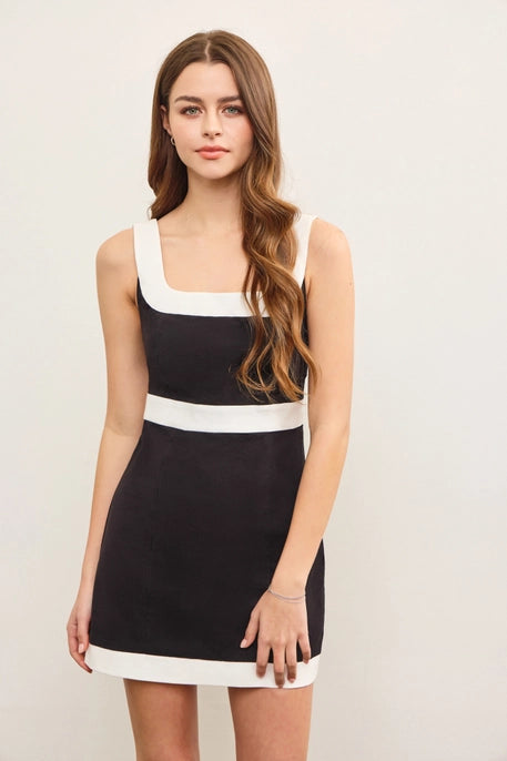 A young woman with long, wavy brown hair wears a black mini dress with a bold white trim along the square neckline, waistline, and hem. The dress has a structured, sleek fit, offering a stylish and modern look against a neutral background.