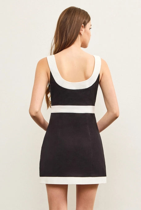 A young woman with long, wavy brown hair wears a black mini dress with a bold white trim along the square neckline, waistline, and hem. The dress has a structured, sleek fit, offering a stylish and modern look against a neutral background.