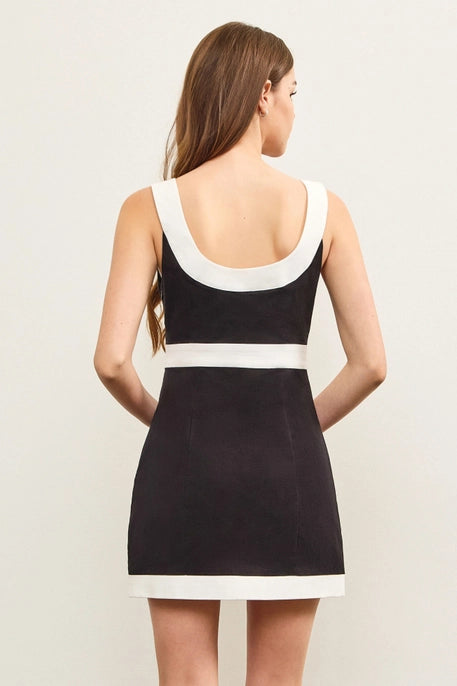 A young woman with long, wavy brown hair wears a black mini dress with a bold white trim along the square neckline, waistline, and hem. The dress has a structured, sleek fit, offering a stylish and modern look against a neutral background.