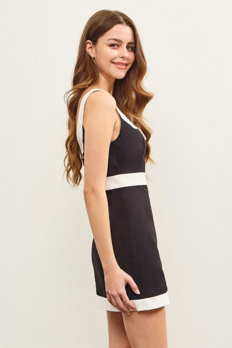 A young woman with long, wavy brown hair wears a black mini dress with a bold white trim along the square neckline, waistline, and hem. The dress has a structured, sleek fit, offering a stylish and modern look against a neutral background.