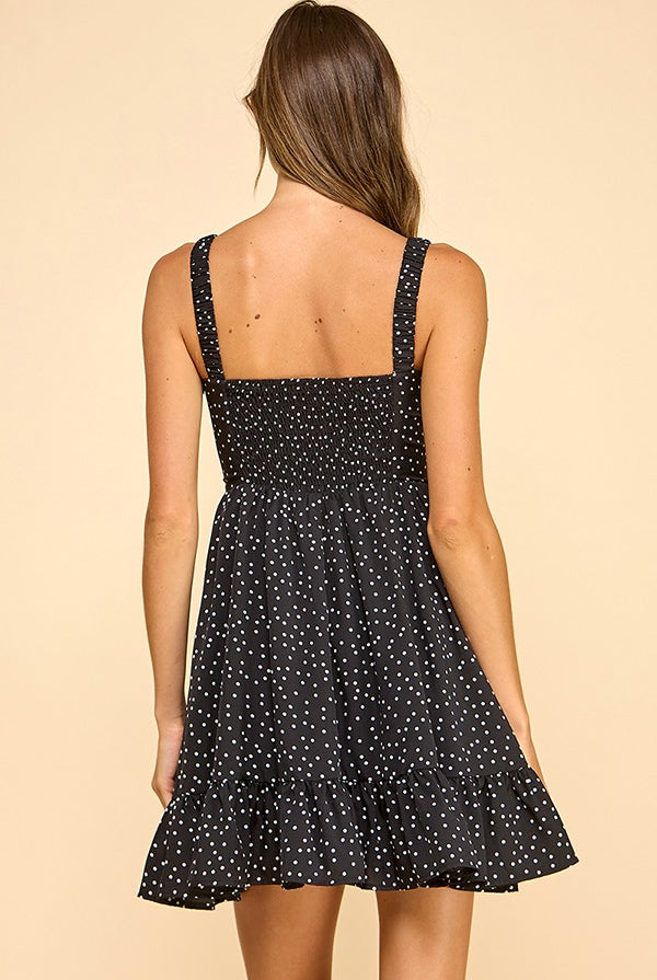 Woman wearing a black mini dress with white polka dots, a sweetheart neckline, and ruffled hem. The dress features wide elastic shoulder straps, a smocked back, and a tiered silhouette for a flattering fit.