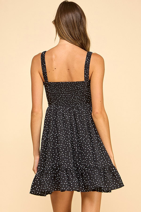 Woman wearing a black mini dress with white polka dots, a sweetheart neckline, and ruffled hem. The dress features wide elastic shoulder straps, a smocked back, and a tiered silhouette for a flattering fit.
