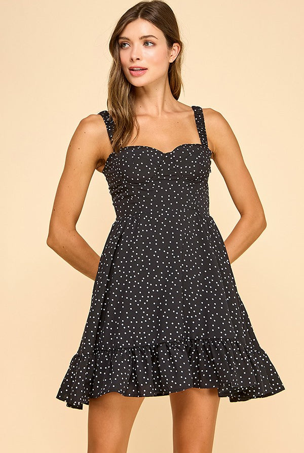 Woman wearing a black mini dress with white polka dots, a sweetheart neckline, and ruffled hem. The dress features wide elastic shoulder straps, a smocked back, and a tiered silhouette for a flattering fit.