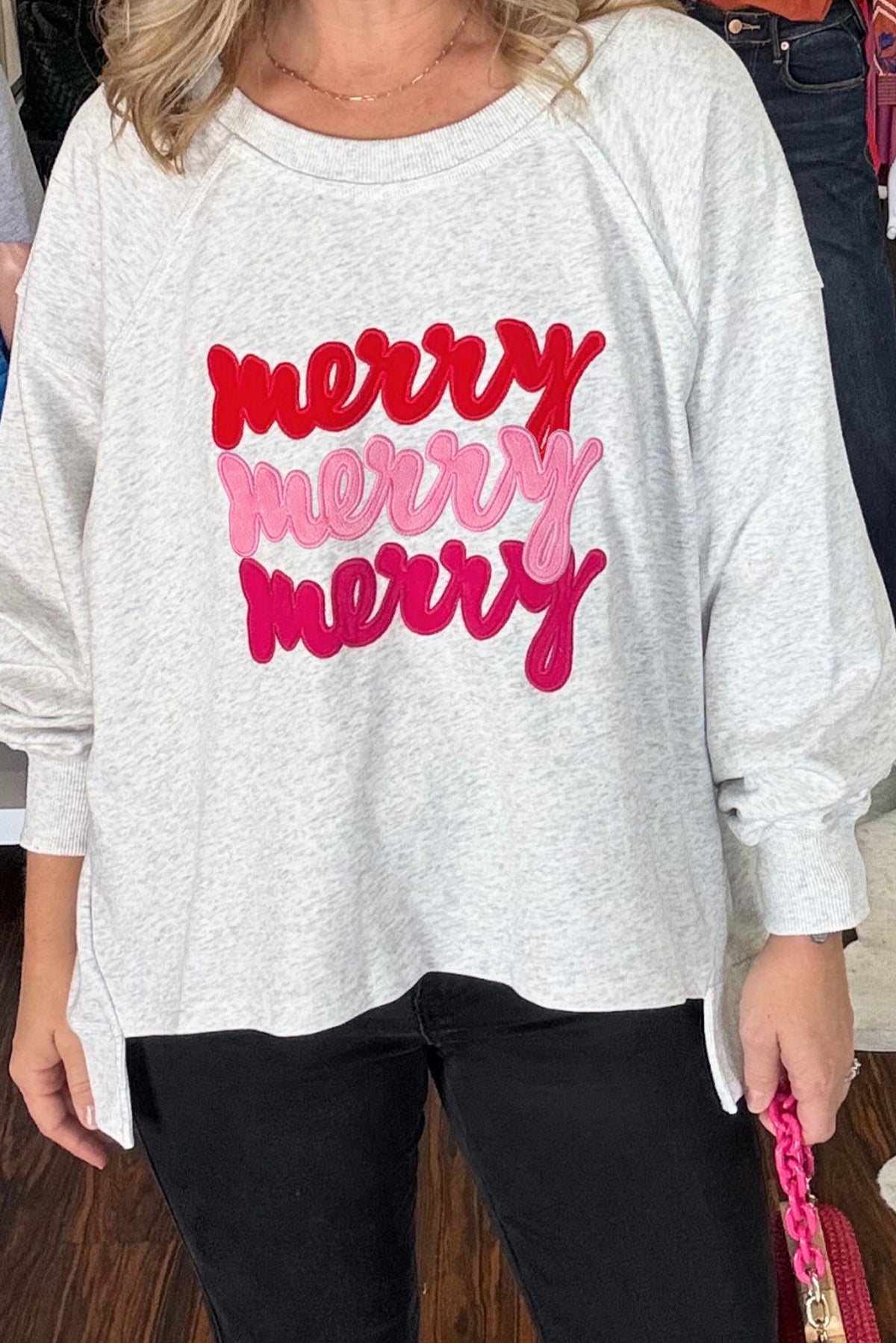 A woman wearing a light heather gray long-sleeve sweatshirt with 'merry merry merry' printed across the front in red, pink, and magenta cursive lettering, paired with black pants.