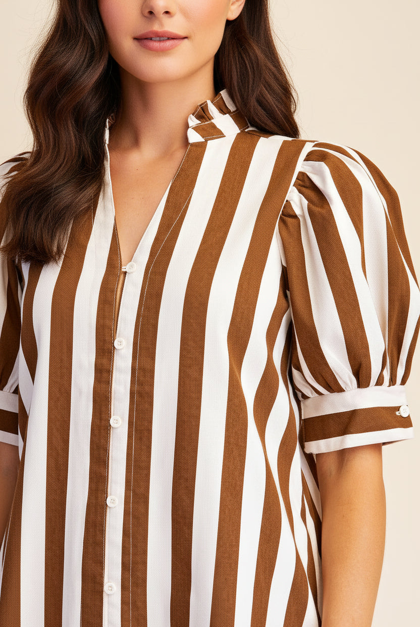 A close-up of a woman wearing a brown and white vertically striped button-up shirt dress with puff sleeves and a tie-neck detail, set against a plain beige background.