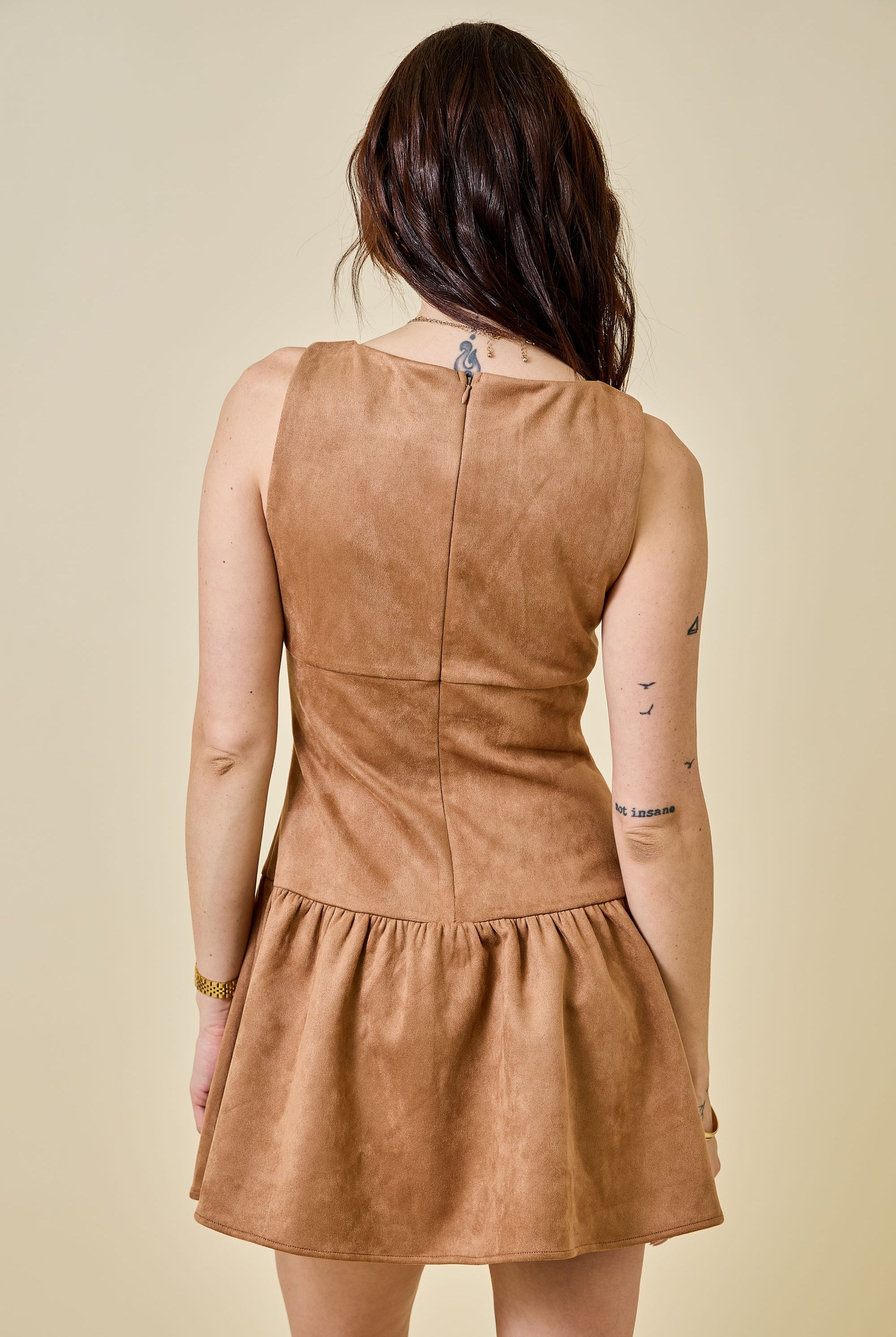 Woman wearing a camel-colored suede mini dress featuring a drop waist with bow ties, sleeveless design, and a back zipper closure. The dress has a soft, structured texture and a flattering A-line silhouette, styled for a chic fall look.