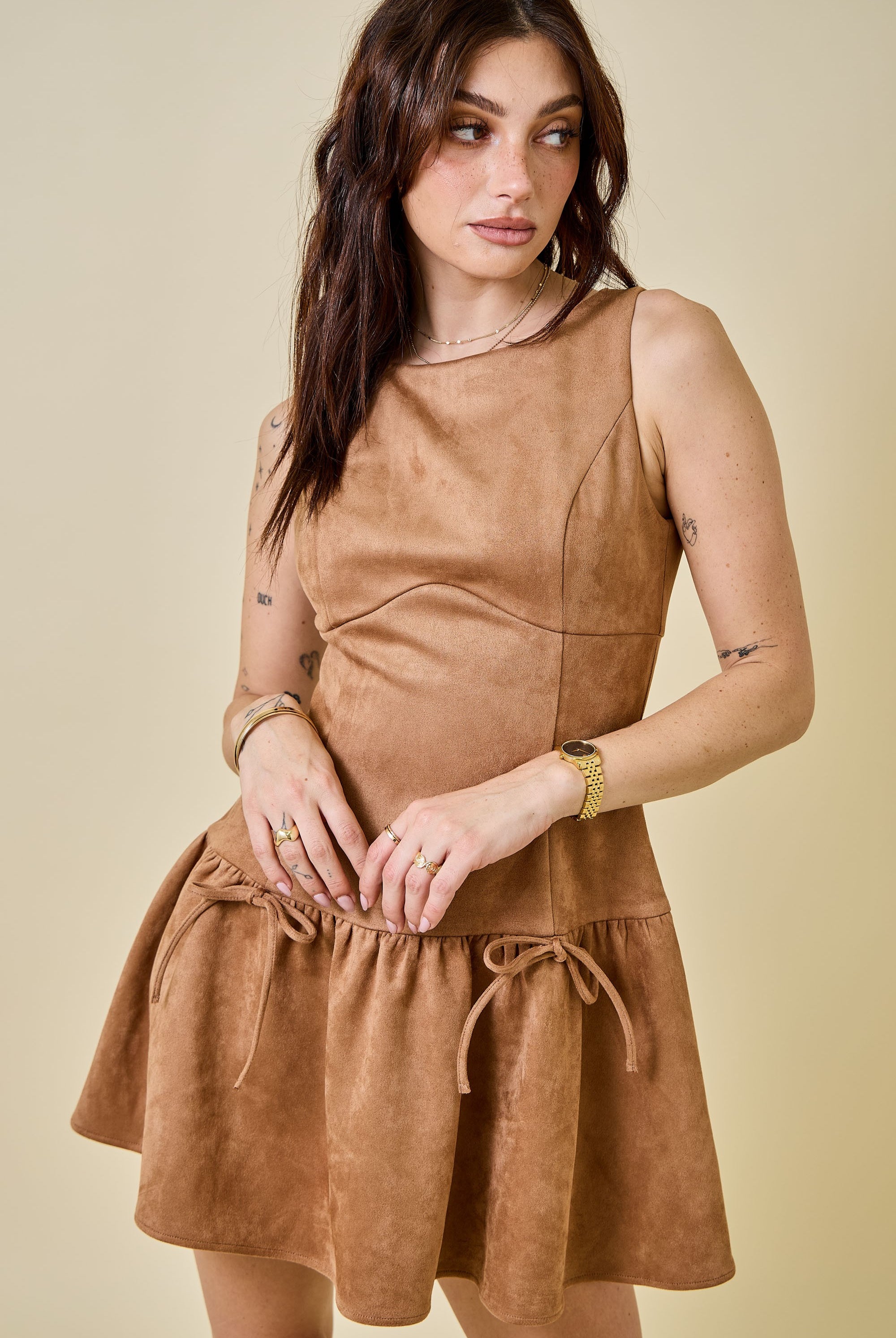 Woman wearing a camel-colored suede mini dress featuring a drop waist with bow ties, sleeveless design, and a back zipper closure. The dress has a soft, structured texture and a flattering A-line silhouette, styled for a chic fall look.