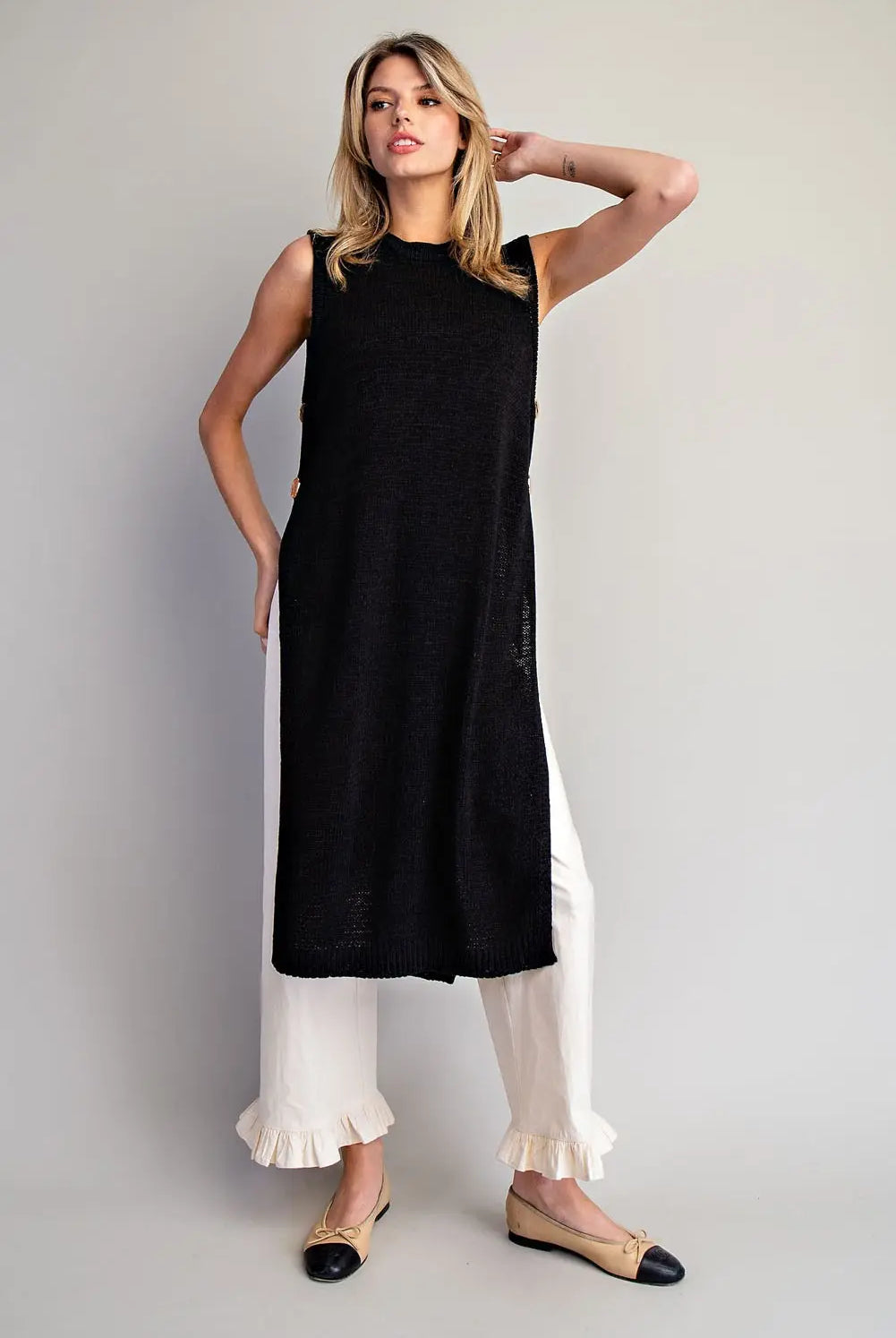 Woman wearing a black sleeveless top and white pants against a gray background
