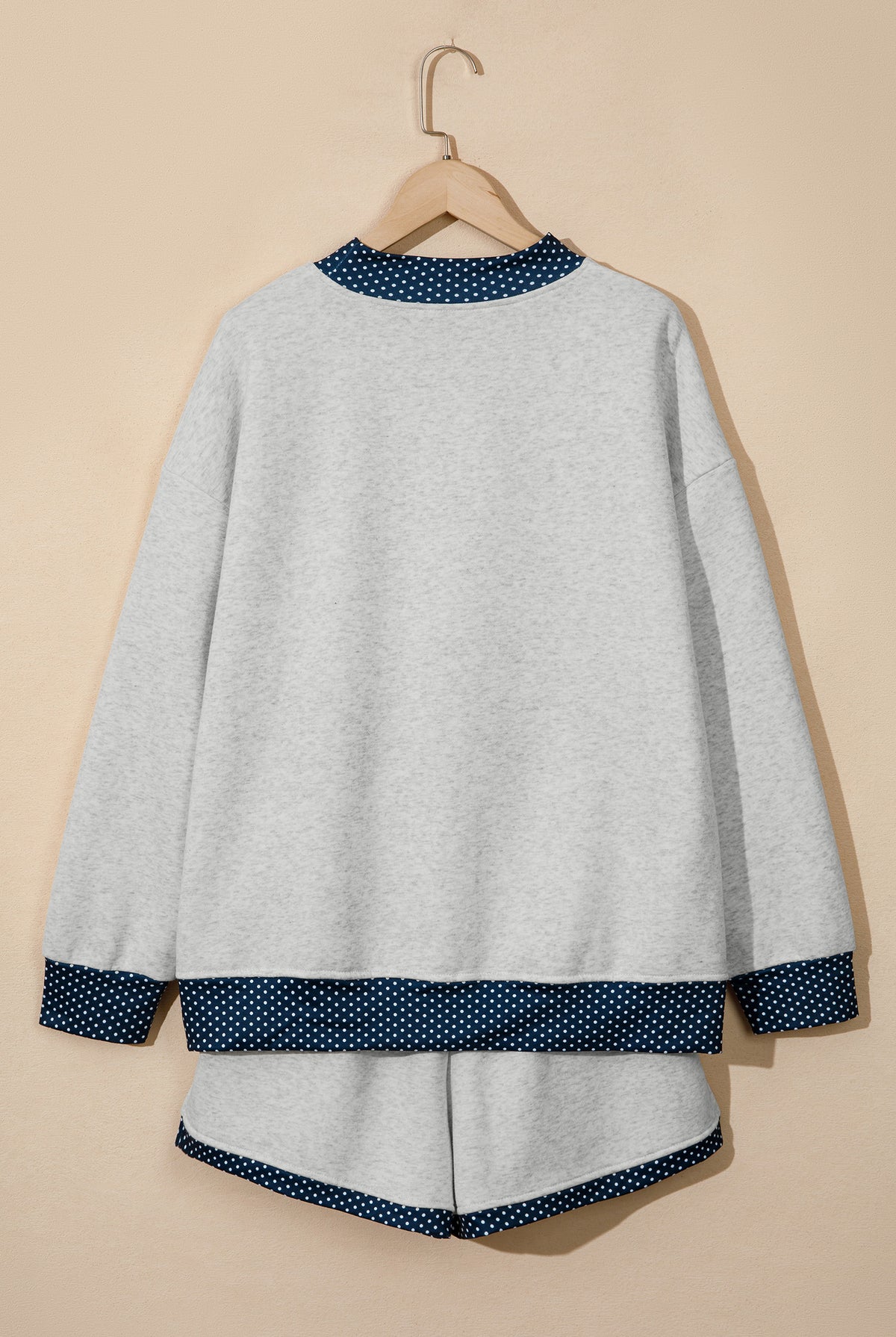 A light gray knit lounge set consisting of a long-sleeve top and matching shorts, hanging on a wooden hanger against a solid beige background. The top and shorts feature contrasting dark blue and white polka dot trim at the collar, cuffs, hem, and bottom of the shorts.