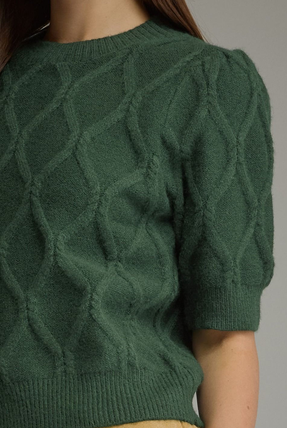 A close-up of a person wearing a dark green short-sleeve cable knit sweater with a textured diamond cable pattern and ribbed collar, cuffs, and hem.