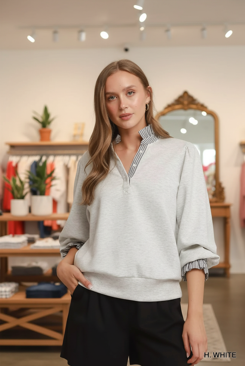 Waist-up front shot of a blonde woman wearing the Umgee heather white sweatshirt top with navy stripe ruffle collar, balloon puff sleeves, and smocked ruffle cuffs, gold hoop earrings, boutique interior