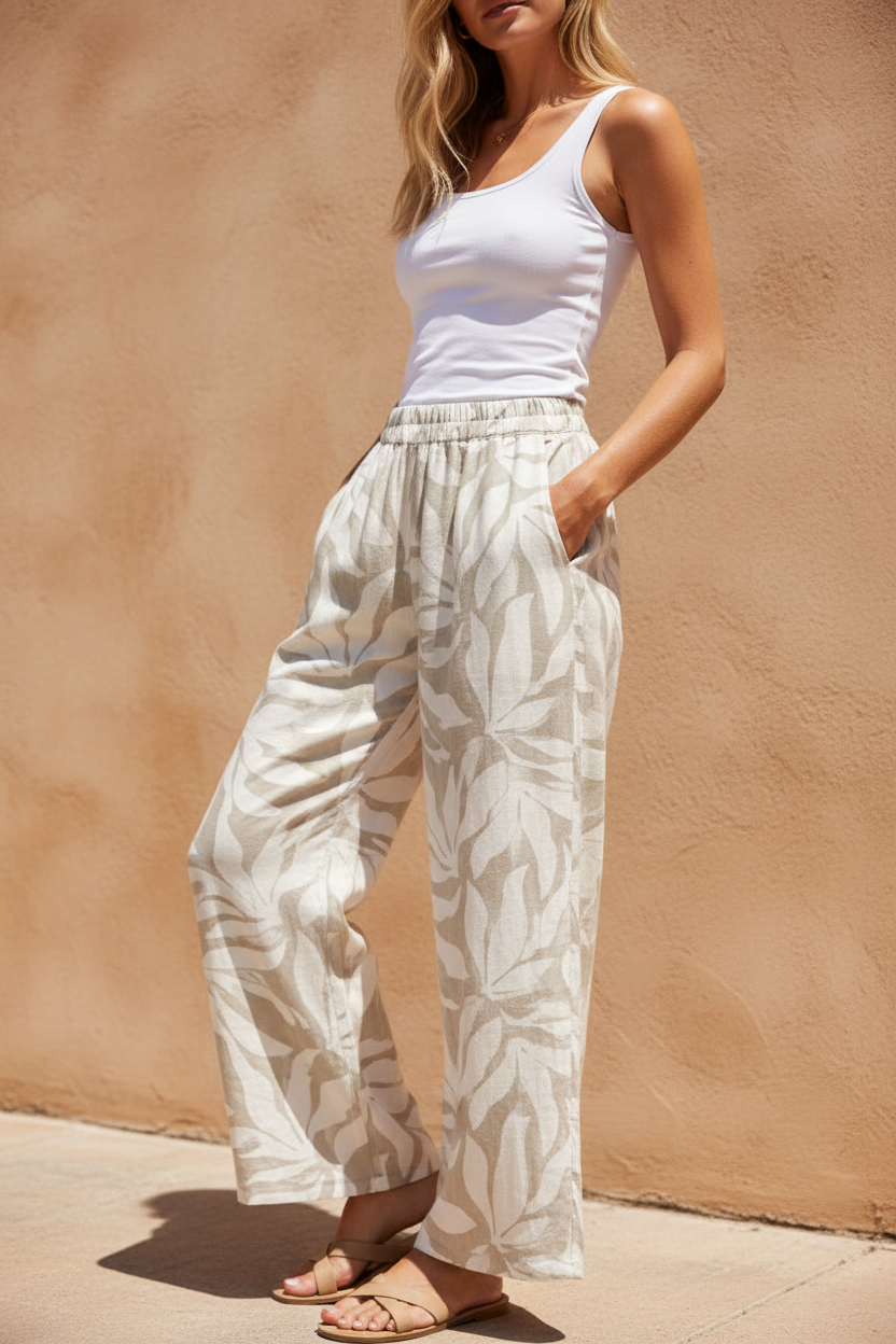 Close-up three-quarter shot of blonde model in taupe botanical print linen wide-leg pants with hands in pockets against stucco wall