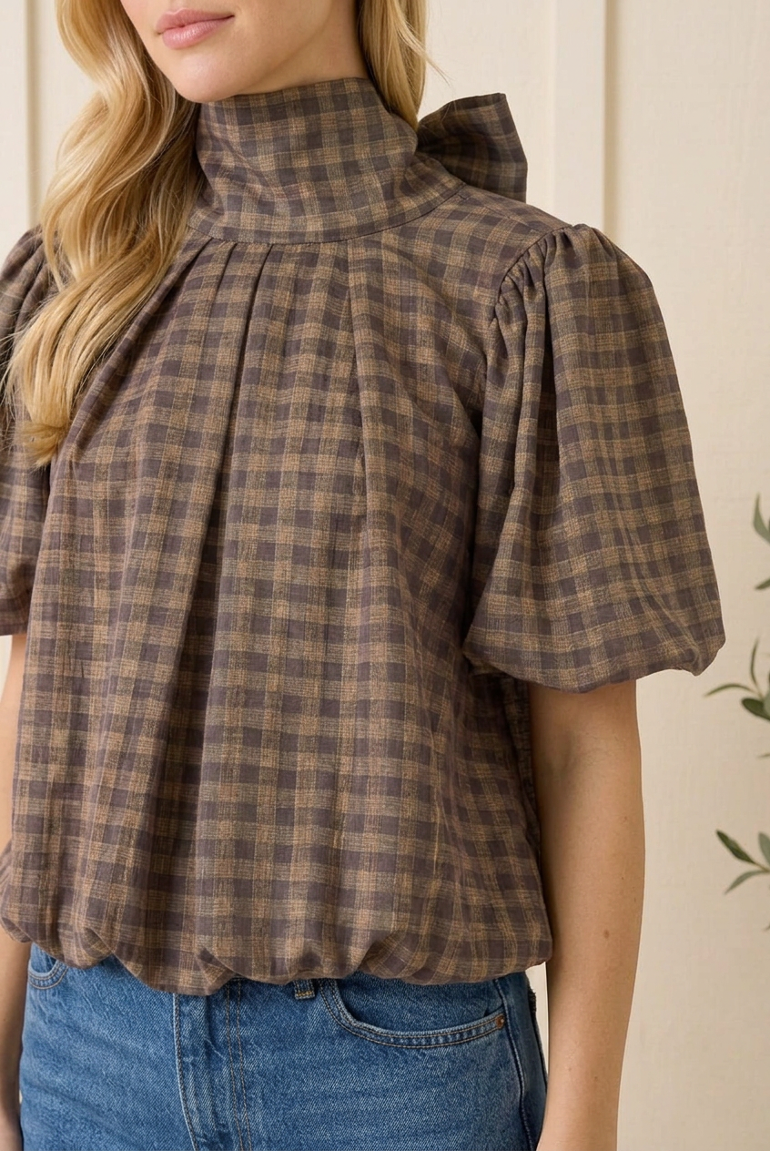 Close-up of brown gingham woven fabric, high neckline, front pleats, puff sleeve and elasticated blouse hem.