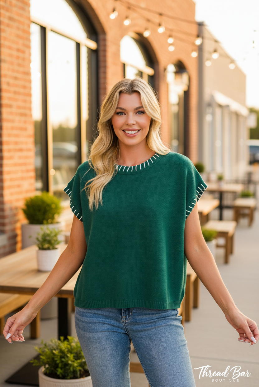 Hunter green short sleeve oversized sweater front view showing white whipstitch detailing along neckline and sleeve edges, relaxed fit styled with denim jeans