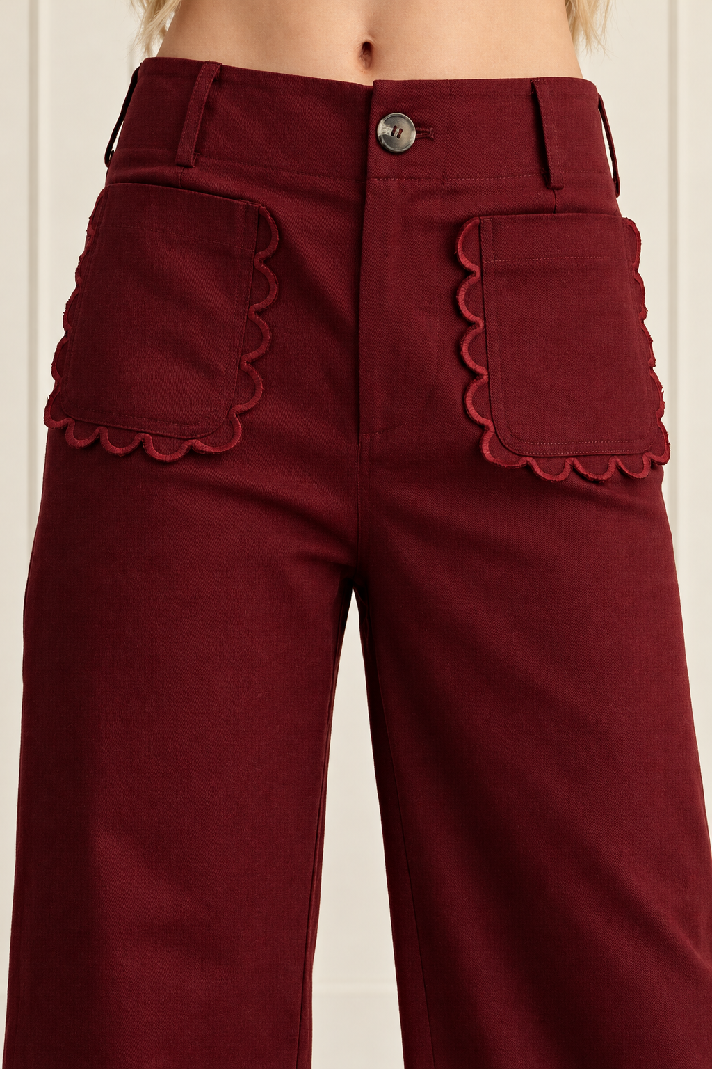 Close-up of burgundy pants highlighting rounded scalloped pocket trim, a metallic waist button and belt loops.