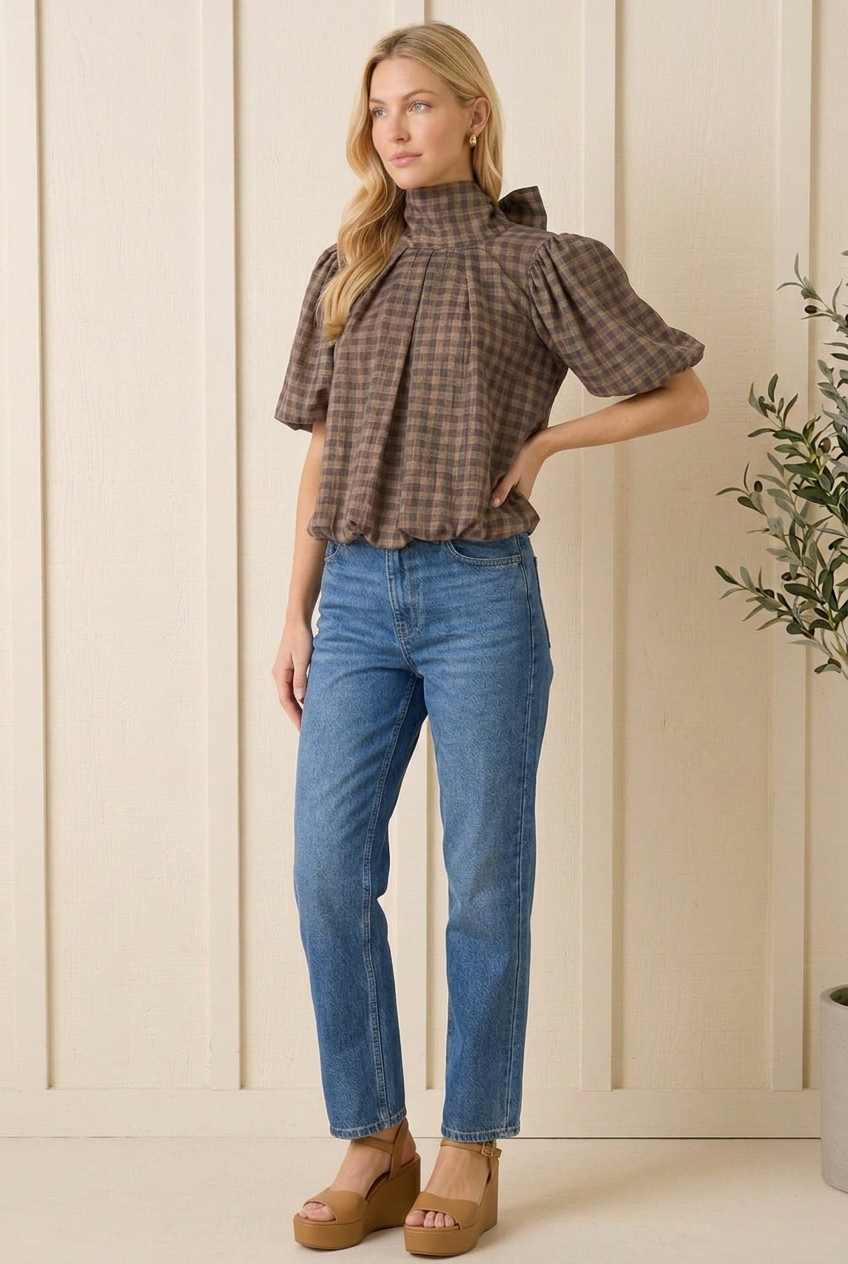 Angled view of brown gingham tie neck blouse showing voluminous short puff sleeves and pleated front.