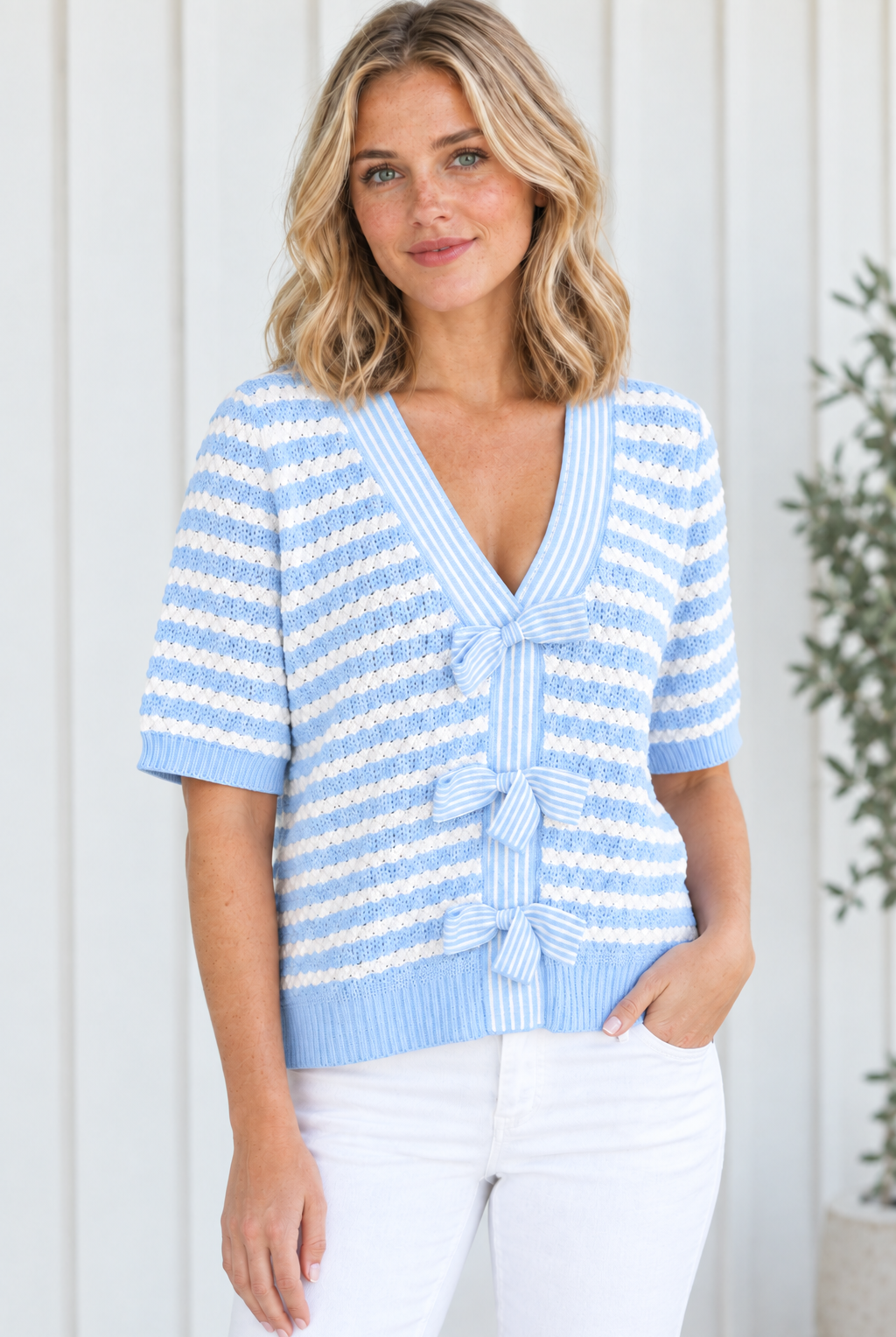 Angled front view of sky blue striped V-neck bow detail knit sweater styled with white jeans