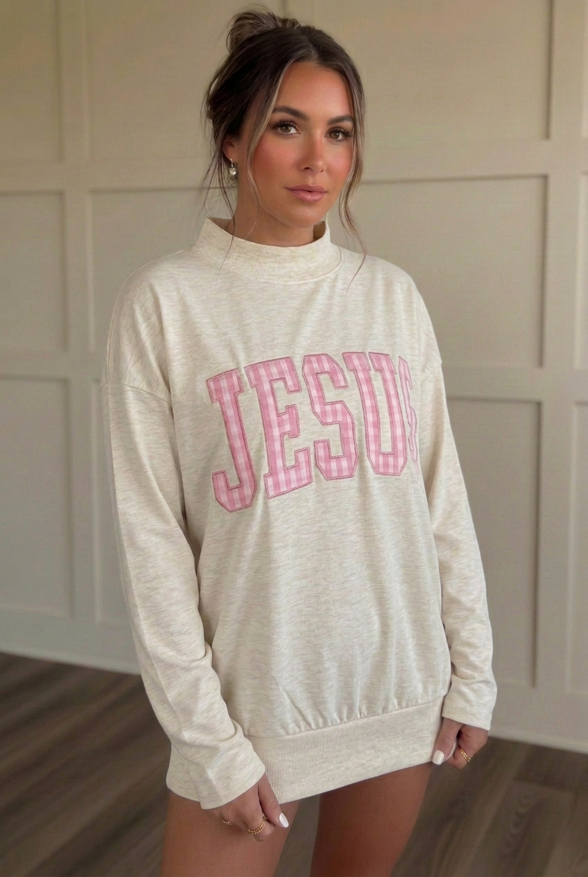 Angled view of cream-gray mock-neck long sleeve tee showing pink gingham JESUS lettering, relaxed sleeves, and a cuffed finished hem.