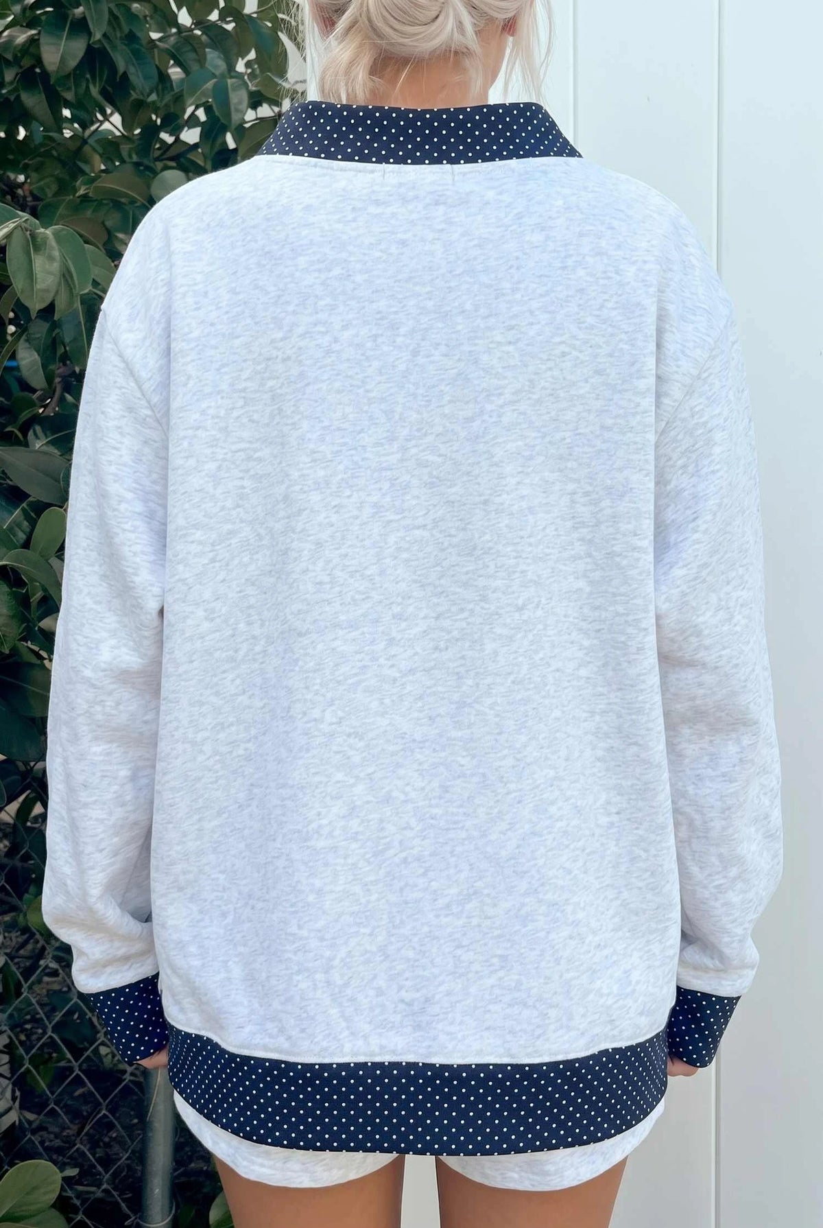 A person shown from the back wearing a light heather gray sweatshirt with dark navy polka-dot trim on the collar, cuffs, and hem, paired with white shorts.