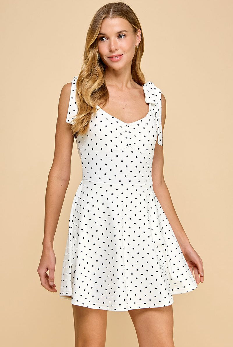 A smiling blonde woman wears a white polka-dotted fit-and-flare dress with bow-tie shoulder straps. The dress features a sweetheart neckline, built-in lined shorts, and a lightweight, breathable fabric, blending feminine style with athleisure-inspired comfort.
