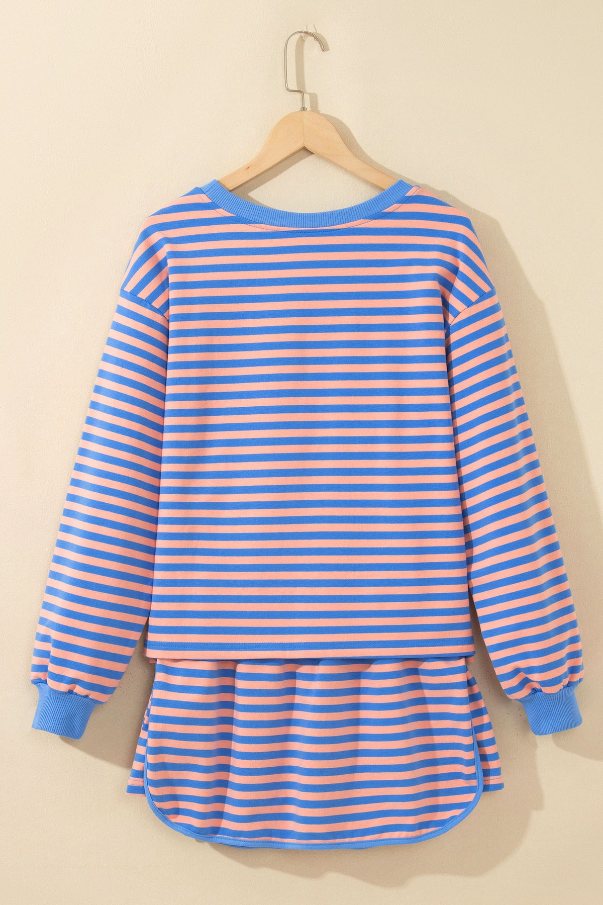 A blue and orange striped long-sleeve tunic top with solid blue ribbed collar, cuffs, and hem, hanging on a wooden hanger against a warm beige wall.