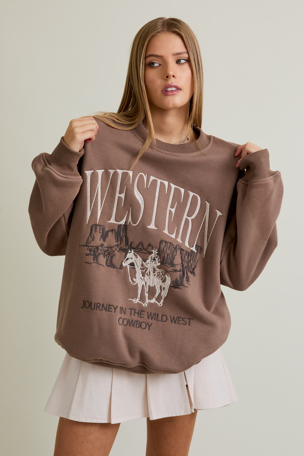 Woman wearing a mocha oversized crewneck sweatshirt with “Western” embroidery and a cowboy graphic
