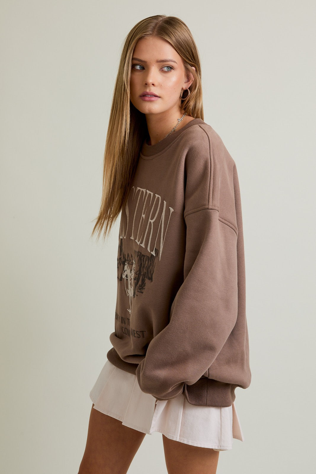 Woman wearing a mocha oversized crewneck sweatshirt with “Western” embroidery and a cowboy graphic