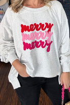 A woman wearing a light heather gray long-sleeve sweatshirt with 'merry merry merry' printed across the front in red, pink, and magenta script, paired with black pants.