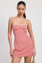 Model wearing a red gingham mini dress with criss-cross tie back, fitted bodice, and adjustable straps, holding a woven handbag — perfect for summer outings or casual date nights.