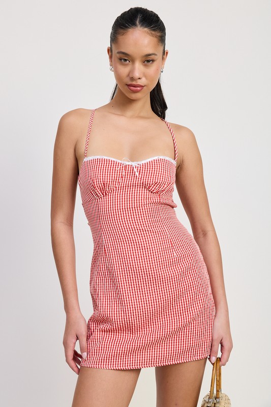 Model wearing a red gingham mini dress with criss-cross tie back, fitted bodice, and adjustable straps, holding a woven handbag — perfect for summer outings or casual date nights.