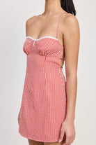 Model wearing a red gingham mini dress with criss-cross tie back, fitted bodice, and adjustable straps, holding a woven handbag — perfect for summer outings or casual date nights.