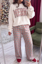 A woman takes a mirror selfie wearing a holiday-themed loungewear set consisting of a cream sweatshirt with a red and green plaid 'MERRY' graphic and matching red, green, and white plaid drawstring pants, paired with red slip-on shoes.