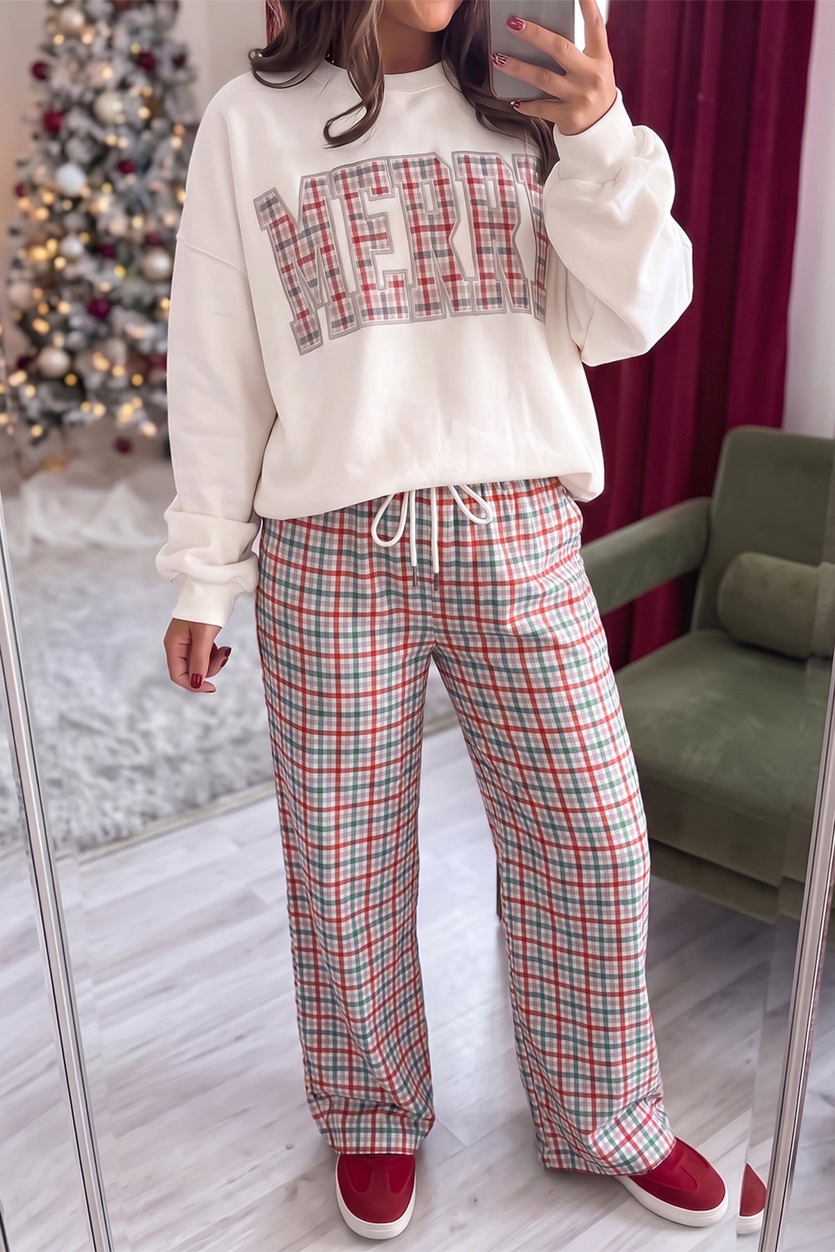 A woman takes a mirror selfie wearing a holiday-themed loungewear set consisting of a cream sweatshirt with a red and green plaid 'MERRY' graphic and matching red, green, and white plaid drawstring pants, paired with red slip-on shoes.