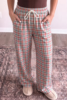A person wearing loose, wide-leg plaid pajama pants with a red, green, and white tartan pattern, a brown top, and beige slippers, standing on a patterned rug.