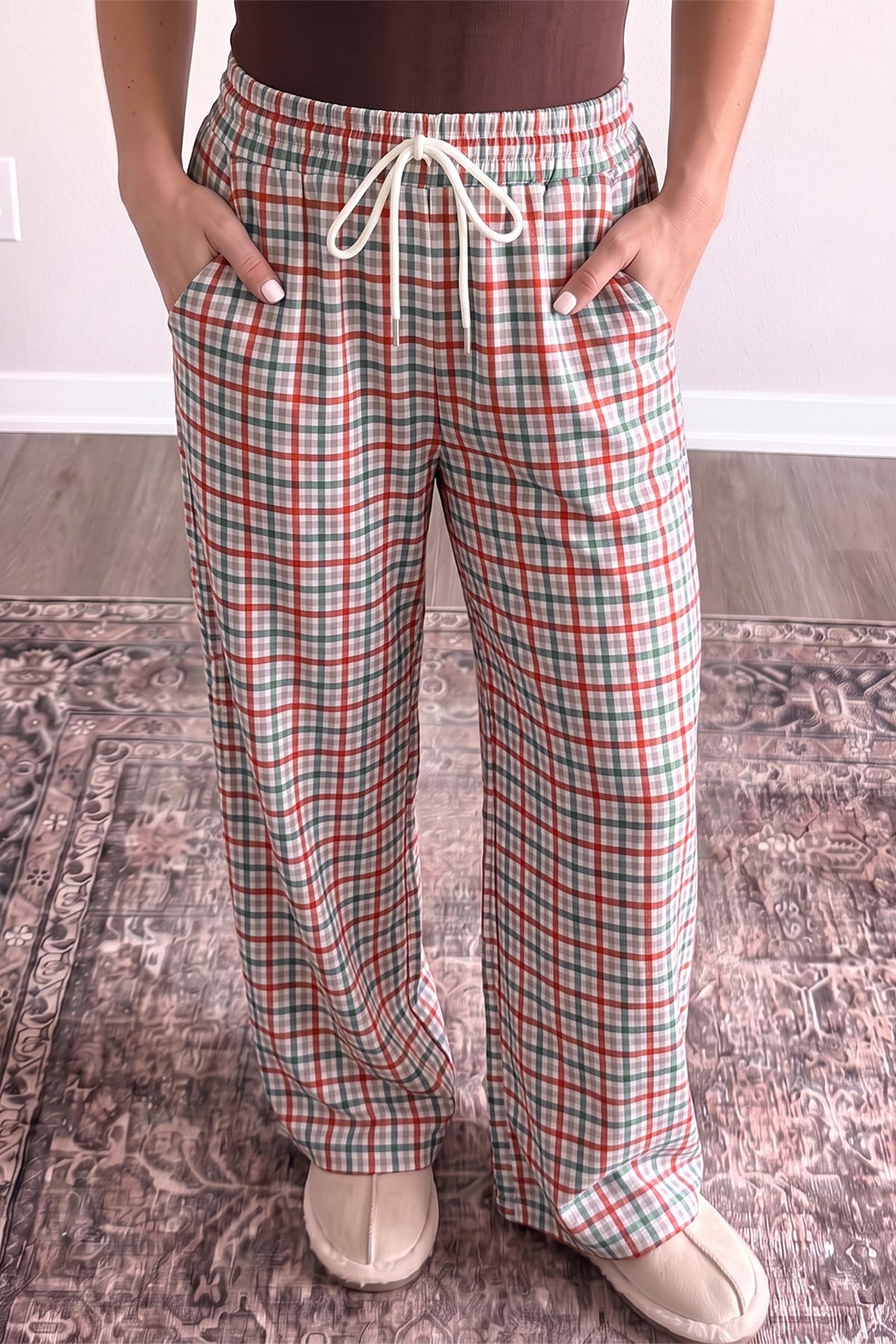 A person wearing loose, wide-leg plaid pajama pants with a red, green, and white tartan pattern, a brown top, and beige slippers, standing on a patterned rug.