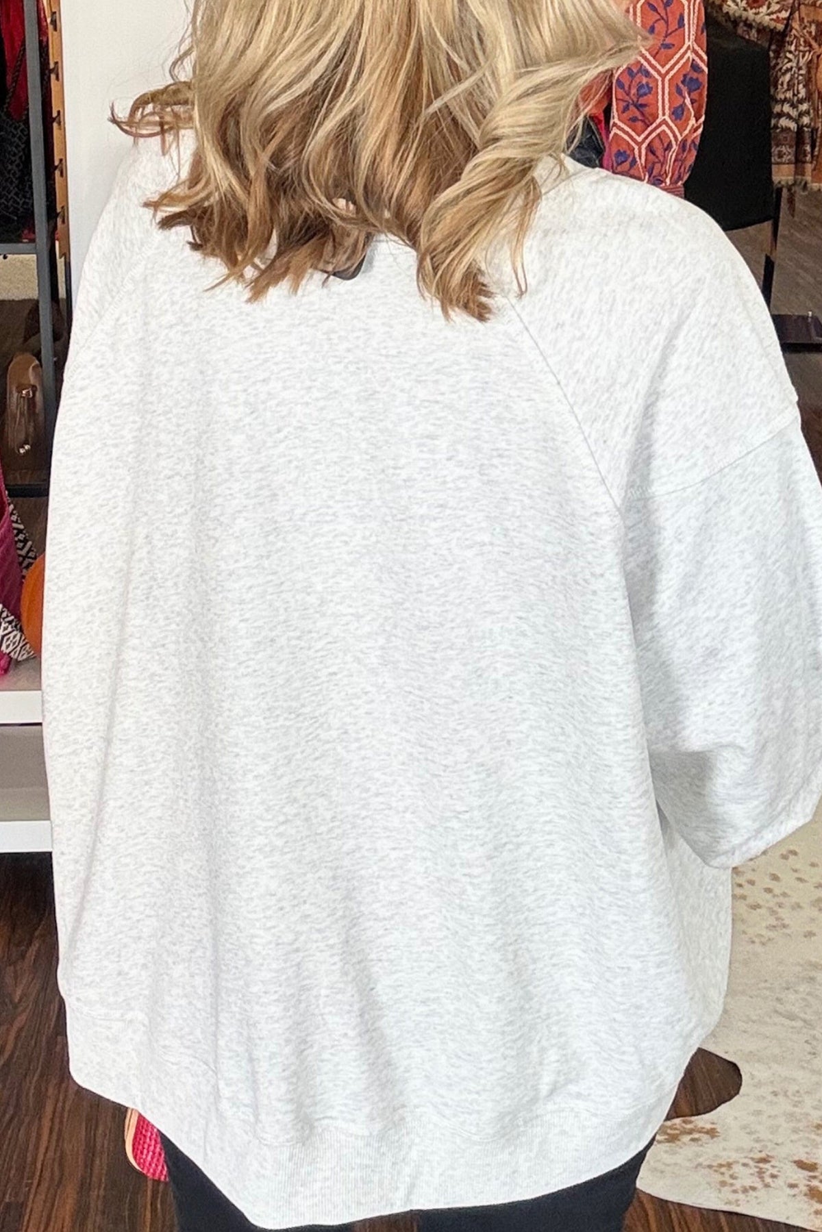 A woman viewed from behind wearing an oversized light heather gray raglan sweatshirt and black pants in a retail store setting.