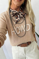 A woman wearing a beige knit sweater with 'HOWDY' in white letters across the chest, styled with a leopard-print neck scarf, layered black chains, a brown tooled belt, and off-white pants.