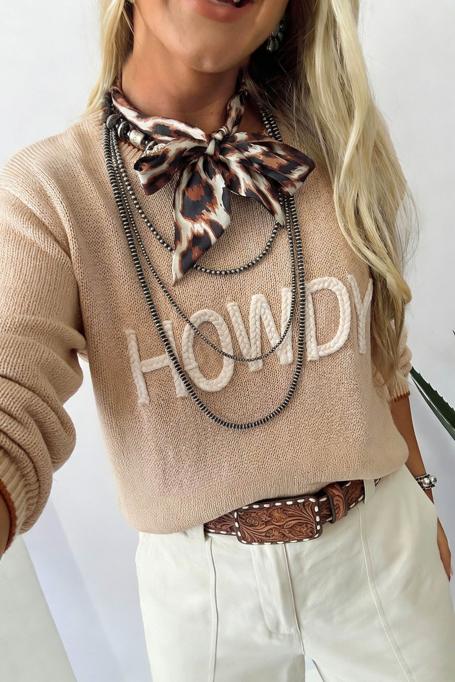 A woman wearing a beige knit sweater with 'HOWDY' in white letters across the chest, styled with a leopard-print neck scarf, layered black chains, a brown tooled belt, and off-white pants.