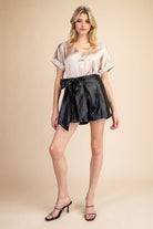 Stylish high-waisted metallic shorts with a satin-like sheen, featuring an oversized bow detail at the waist. Perfect for night-out outfits, party looks, and trendy fashion styles. Model wearing black shorts with bow tie