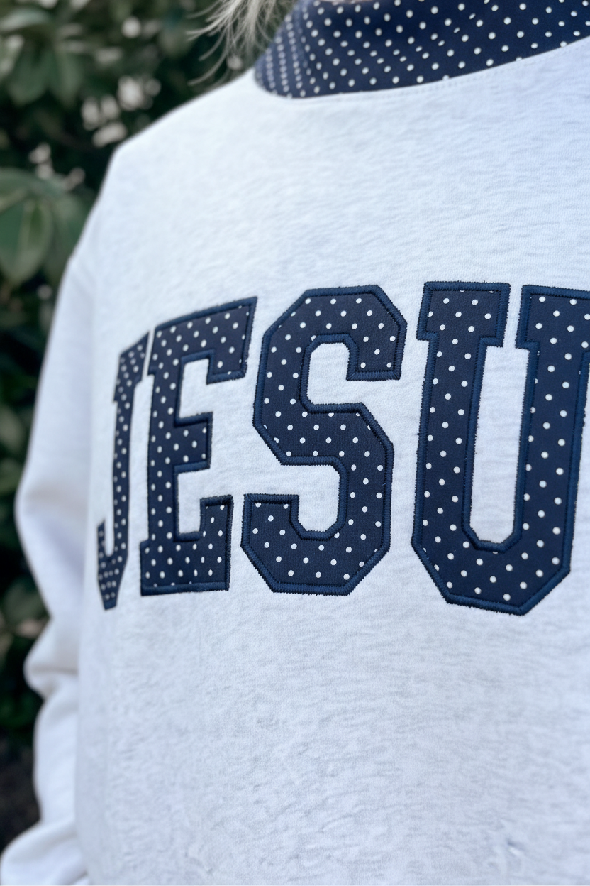 Close-up of bold JESUS navy polka dot appliqé lettering on light heather grey oversized mock neck sweatshirt — collegiate style letters with white polka dot fabric detail and navy contrast collar trim