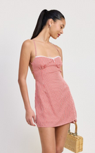 Model wearing a red gingham mini dress with criss-cross tie back, fitted bodice, and adjustable straps, holding a woven handbag — perfect for summer outings or casual date nights.