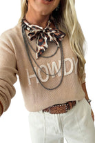 A woman wearing a beige knit sweater with 'HOWDY' in white letters, styled with a leopard-print neck scarf, layered black chains, a brown tooled belt, and white pants against a white background.