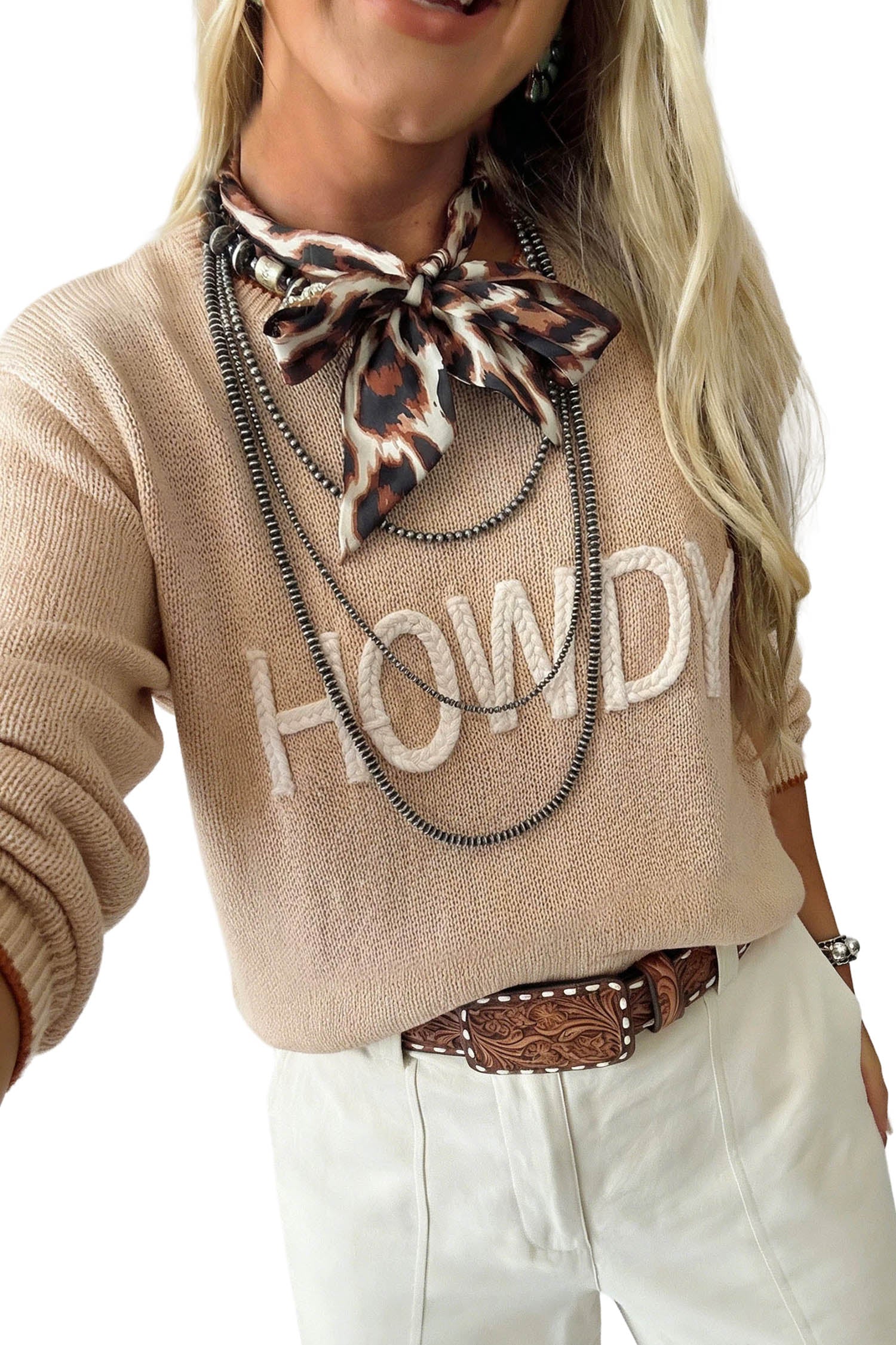 A woman wearing a beige knit sweater with 'HOWDY' in white letters, styled with a leopard-print neck scarf, layered black chains, a brown tooled belt, and white pants against a white background.
