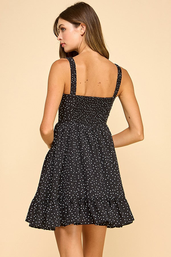 Woman wearing a black mini dress with white polka dots, a sweetheart neckline, and ruffled hem. The dress features wide elastic shoulder straps, a smocked back, and a tiered silhouette for a flattering fit.